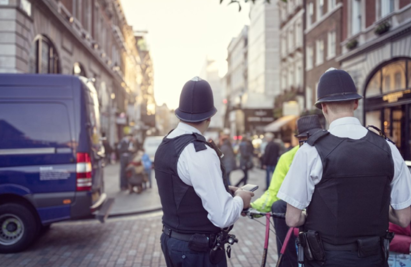 Two police officers in uniform stand on a busy city street near a police van, while pedestrians walk along the cobblestone road—capturing a typical day in the Westminster City Council area.