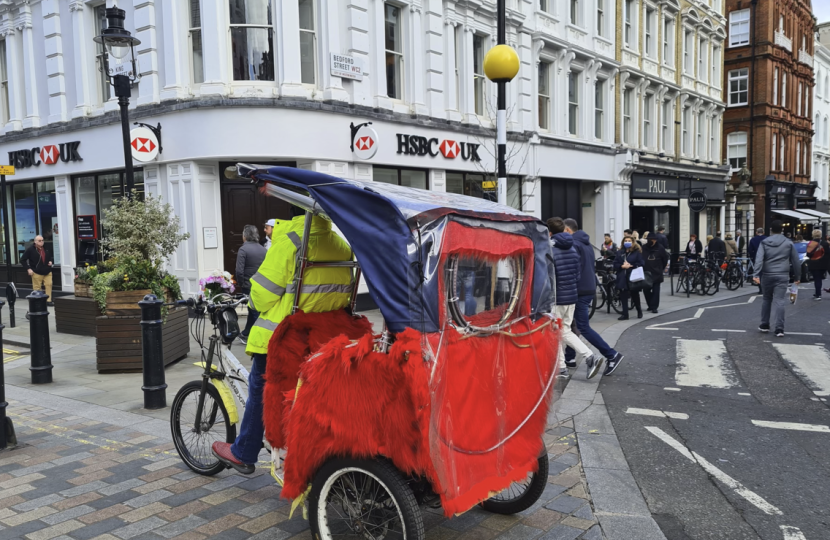 A person wearing a yellow jacket rides a red and blue rickshaw on a city street, with people walking on the pavements and white Westminster buildings in the background