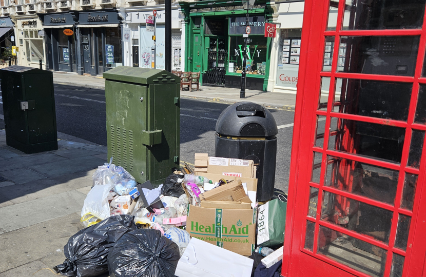 A pile of rubbish bags, cardboard, and other waste is left on the pavement next to a red phone box on a city street