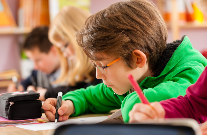 A young boy in a green hoodie writes with a pencil at a desk in a classroom. Other students are seated beside him, also focused on their work. The softly blurred background hints at an engaging learning environment