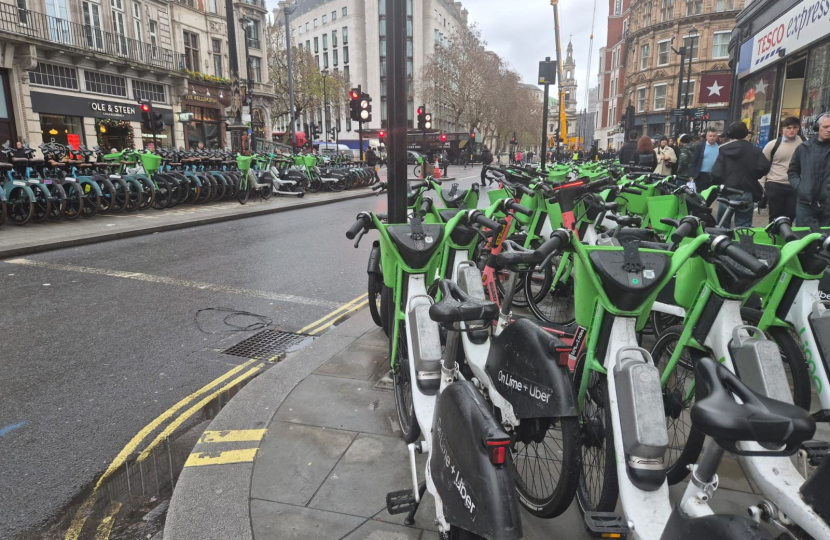 A row of green rental bikes is parked blocking a pavement, with cars visible on the road and people walking by. Buildings line both sides of the street under cloudy skies