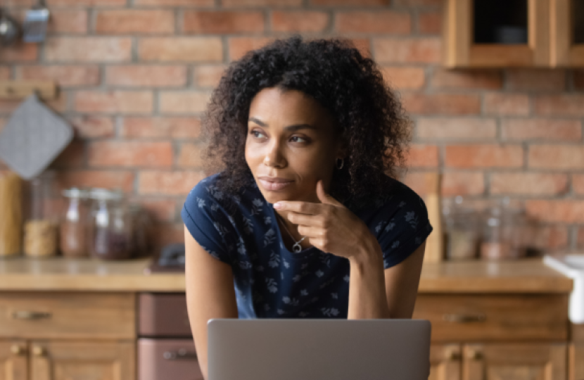 A woman with curly hair, wearing a navy polka dot shirt, rests her chin on her hand while looking thoughtfully to the side. She sits in a kitchen with a laptop
