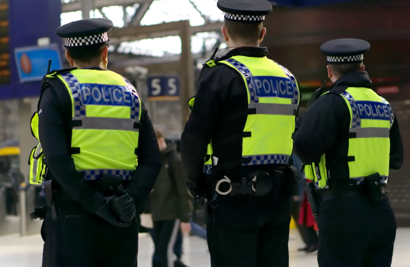 Three police officers in high-visibility jackets stand in a City of London train station, facing away from the camera. There are people and a digital departure board in the background.