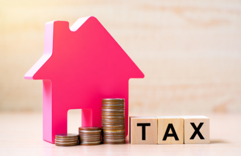 A pink house-shaped figure, stacks of coins, and wooden blocks spelling “TAX” sit on a wooden surface, symbolizing property or real estate taxes