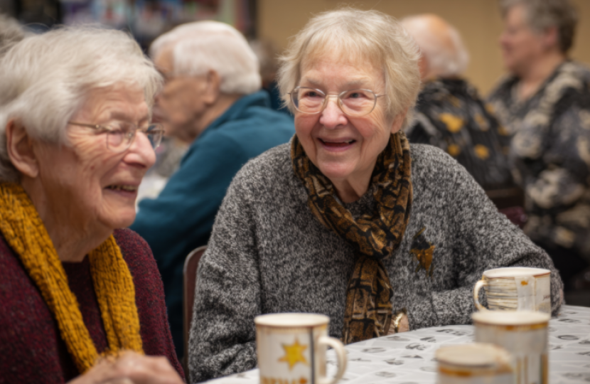 Two elderly women sit at a table, smiling and chatting over mugs, perhaps discussing the latest news. Other seniors gather in the background, suggesting a social event in a warm indoor setting.