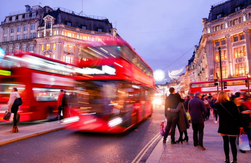 Busy Oxford Street at dusk with blurred red double-decker buses passing by people walking on the pavement, and historic buildings aglow