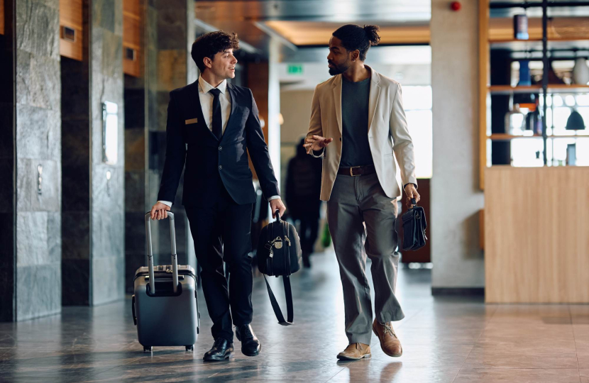Two people in business attire walk indoors with rolling suitcases. One wears a black suit and the other a light blazer with a gray shirt and pants. They appear to be conversing while moving through a modern building.