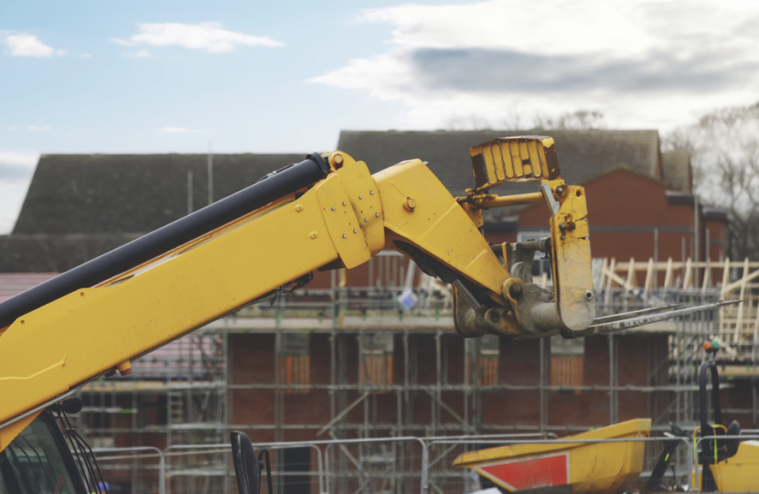 A yellow construction vehicle extends its arm toward a partially built structure with scaffolding, as two workers in safety gear stand on the upper level. Houses and a cloudy sky are visible in the background.