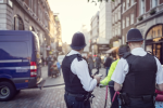 Two police officers in uniform stand on a busy city street near a police van, while pedestrians walk along the cobblestone road—capturing a typical day in the Westminster City Council area.