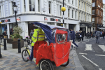 A person wearing a yellow jacket rides a red and blue rickshaw on a city street, with people walking on the pavements and white Westminster buildings in the background