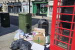 A pile of rubbish bags, cardboard, and other waste is left on the pavement next to a red phone box on a city street