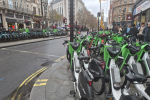 A row of green rental bikes is parked blocking a pavement, with cars visible on the road and people walking by. Buildings line both sides of the street under cloudy skies