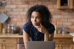 A woman with curly hair, wearing a navy polka dot shirt, rests her chin on her hand while looking thoughtfully to the side. She sits in a kitchen with a laptop