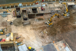 A construction site with several excavators digging, materials scattered around, unfinished concrete structures, and workers in safety gear operating equipment