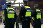 Three police officers in high-visibility jackets stand in a City of London train station, facing away from the camera. There are people and a digital departure board in the background.