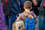 A young boy with short brown hair, wearing a brown hoodie and a blue backpack holding a stuffed horse toy, stands among a group of people with backpacks, possibly waiting for transport