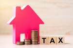 A pink house-shaped figure, stacks of coins, and wooden blocks spelling “TAX” sit on a wooden surface, symbolizing property or real estate taxes