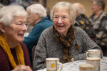 Two elderly women sit at a table, smiling and chatting over mugs, perhaps discussing the latest news. Other seniors gather in the background, suggesting a social event in a warm indoor setting.
