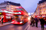 Busy Oxford Street at dusk with blurred red double-decker buses passing by people walking on the pavement, and historic buildings aglow