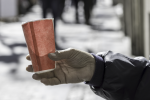 A close-up photograph captures a hand holding a small, paper cup with a hot beverage outdoors on a cold winter day, emphasizing the serenity and quiet of a chilly moment.  The scene composition centers on the hand and the cup, which are positioned in the foreground. The hand, clad in a dark jacket with a visible cuff, grips the cup gently, with fingers wrapped around its textured surface. The background is blurred, featuring snow-covered ground and indistinct figures or objects
