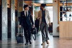 Two people in business attire walk indoors with rolling suitcases. One wears a black suit and the other a light blazer with a gray shirt and pants. They appear to be conversing while moving through a modern building.