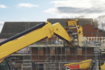 A yellow construction vehicle extends its arm toward a partially built structure with scaffolding, as two workers in safety gear stand on the upper level. Houses and a cloudy sky are visible in the background.