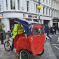 A person wearing a yellow jacket rides a red and blue rickshaw on a city street, with people walking on the pavements and white Westminster buildings in the background