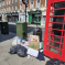 A pile of rubbish bags, cardboard, and other waste is left on the pavement next to a red phone box on a city street