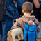 A young boy with short brown hair, wearing a brown hoodie and a blue backpack holding a stuffed horse toy, stands among a group of people with backpacks, possibly waiting for transport