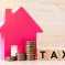 A pink house-shaped figure, stacks of coins, and wooden blocks spelling “TAX” sit on a wooden surface, symbolizing property or real estate taxes