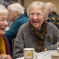 Two elderly women sit at a table, smiling and chatting over mugs, perhaps discussing the latest news. Other seniors gather in the background, suggesting a social event in a warm indoor setting.