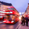 Busy Oxford Street at dusk with blurred red double-decker buses passing by people walking on the pavement, and historic buildings aglow
