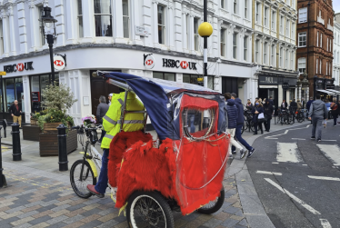 A person wearing a yellow jacket rides a red and blue rickshaw on a city street, with people walking on the pavements and white Westminster buildings in the background