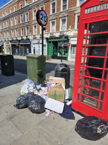 A pile of rubbish bags, cardboard, and other waste is left on the pavement next to a red phone box on a city street