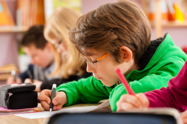 A young boy in a green hoodie writes with a pencil at a desk in a classroom. Other students are seated beside him, also focused on their work. The softly blurred background hints at an engaging learning environment