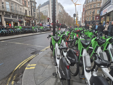 A row of green rental bikes is parked blocking a pavement, with cars visible on the road and people walking by. Buildings line both sides of the street under cloudy skies