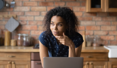 A woman with curly hair, wearing a navy polka dot shirt, rests her chin on her hand while looking thoughtfully to the side. She sits in a kitchen with a laptop
