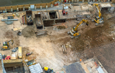 A construction site with several excavators digging, materials scattered around, unfinished concrete structures, and workers in safety gear operating equipment
