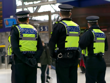 Three police officers in high-visibility jackets stand in a City of London train station, facing away from the camera. There are people and a digital departure board in the background.