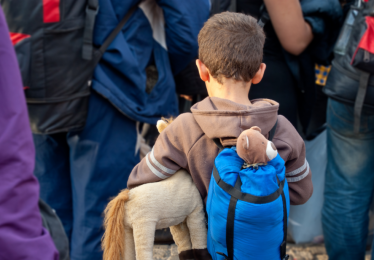 A young boy with short brown hair, wearing a brown hoodie and a blue backpack holding a stuffed horse toy, stands among a group of people with backpacks, possibly waiting for transport