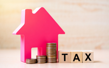 A pink house-shaped figure, stacks of coins, and wooden blocks spelling “TAX” sit on a wooden surface, symbolizing property or real estate taxes