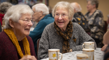 Two elderly women sit at a table, smiling and chatting over mugs, perhaps discussing the latest news. Other seniors gather in the background, suggesting a social event in a warm indoor setting.
