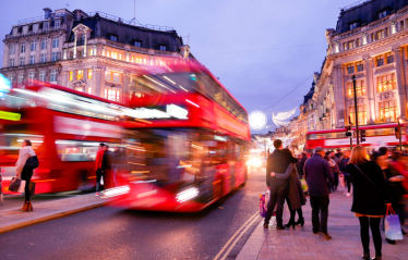 Busy Oxford Street at dusk with blurred red double-decker buses passing by people walking on the pavement, and historic buildings aglow
