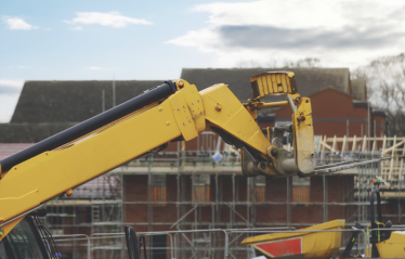 A yellow construction vehicle extends its arm toward a partially built structure with scaffolding, as two workers in safety gear stand on the upper level. Houses and a cloudy sky are visible in the background.
